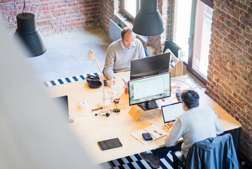 Two professionals collaborating in a modern workspace, featuring multiple computer screens, a wooden table, and a brick wall backdrop, representing a productive environment for marketing automation solutions in Connecticut.