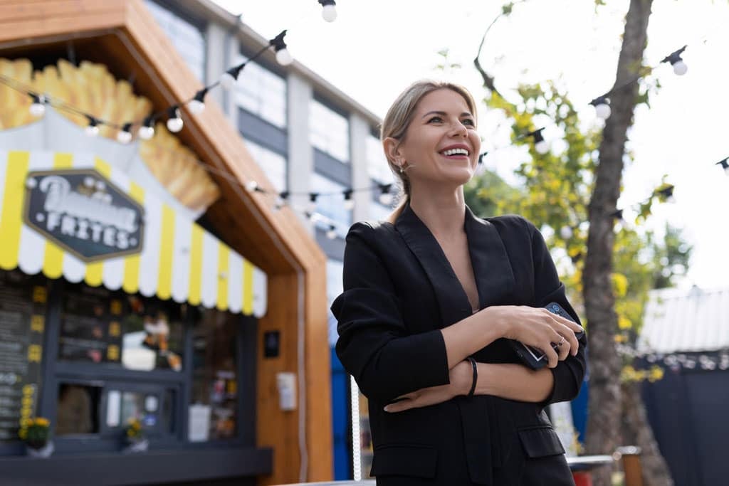 Smiling businesswoman in a black suit standing outside a street café with a vibrant yellow-striped awning, reflecting Connecticut's lively business atmosphere.